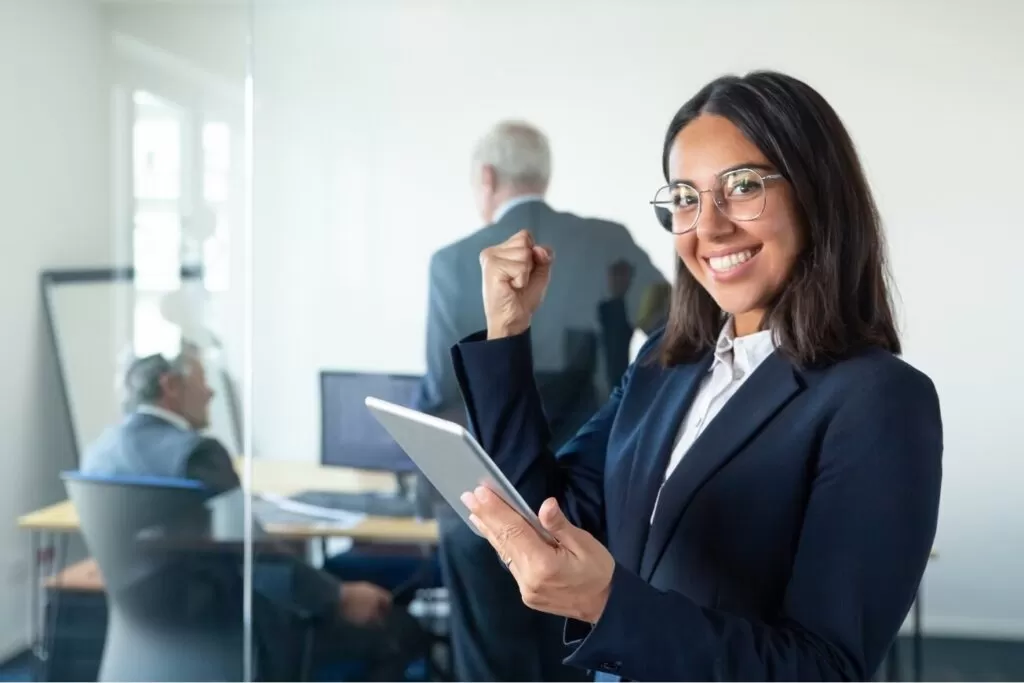 happy-female-professional-glasses-suit-holding-tablet-making-winner-gesture-while-two-businessmen-working-glass-wall-copy-space-communication-concept (1)